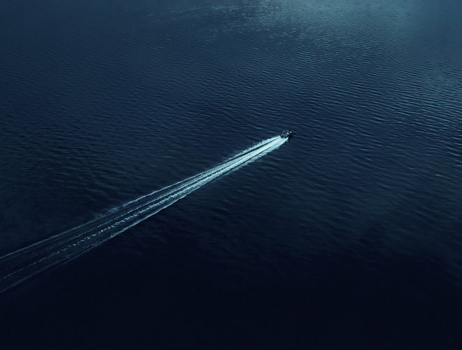 Aerial view of a small motorboat cutting diagonally across deep blue water, leaving a long white wake behind it.
