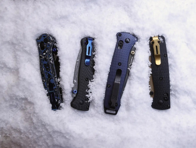 Four folding knives with blue, black, and gold accents lie partially buried in fresh white snow.