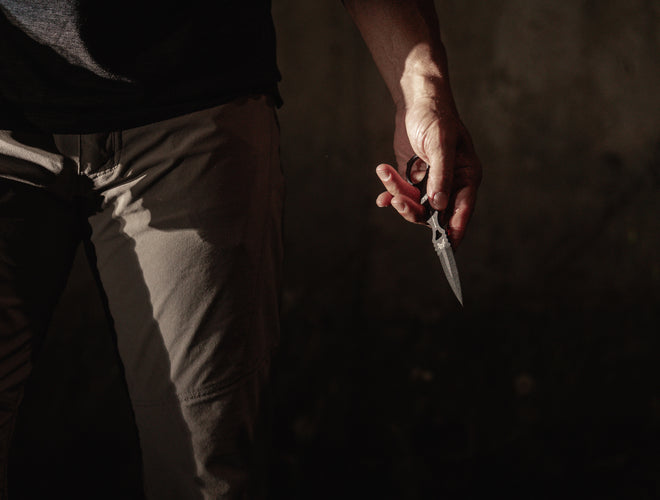 Person's hand holds small silver knife against dark pants in dramatic, shadowy lighting.
