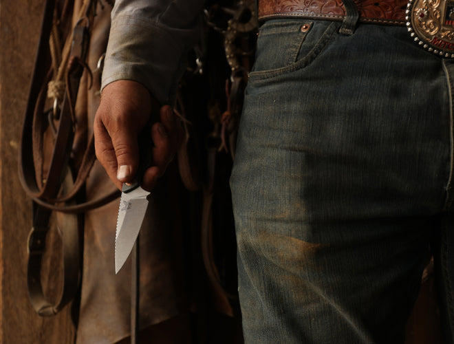Person in jeans and ornate belt holds a serrated knife near hanging leather tack in dim, warm light.