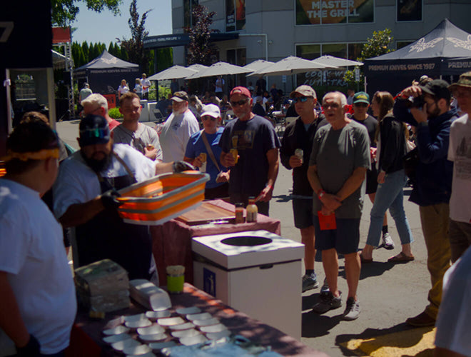 Crowd watches bearded chef pour sauce at sunny outdoor food event with tents and "Master the Grill" sign.