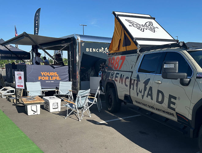 Benchmade branded truck with rooftop tent parked beside a trailer and camp chairs under blue sky.