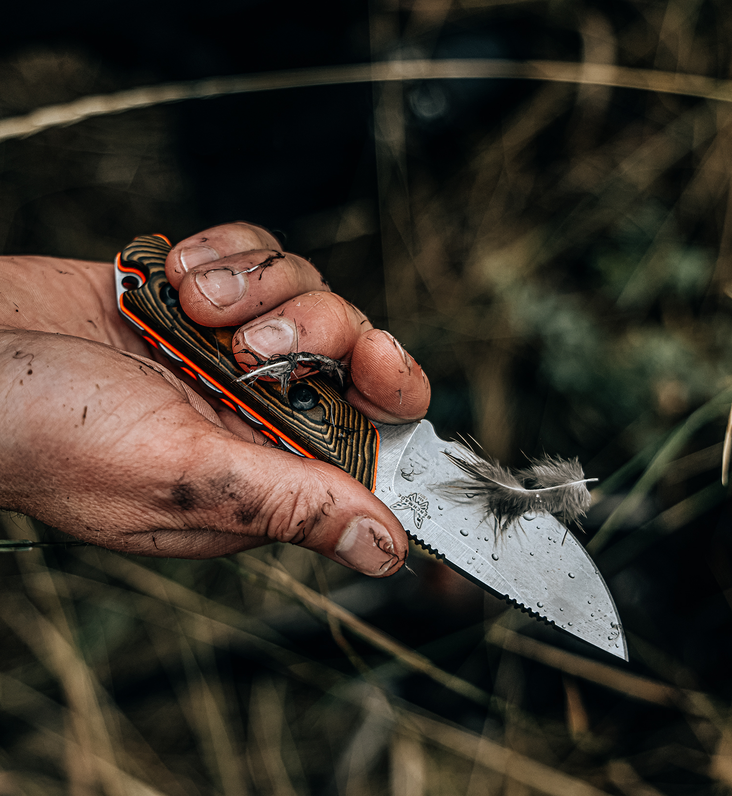 Dirty hand grips orange-accented knife with feather on blade against blurred grassy background.