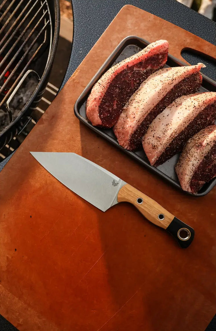 Wooden-handled knife on leather board beside seasoned raw steaks, with charcoal grill visible in background.