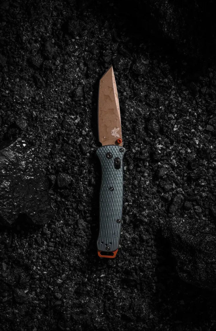 Folding knife with bronze blade and textured blue-gray handle rests on dark, wet gravel.