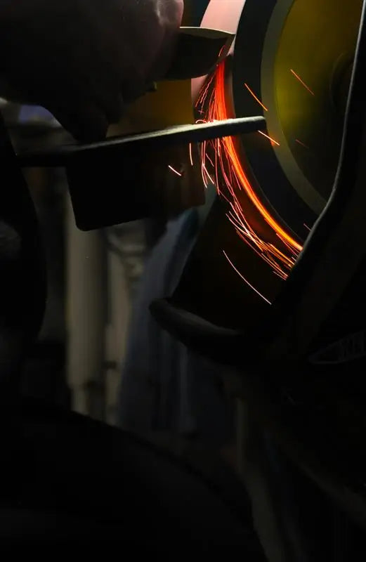 Dark close-up of metal grinding on a wheel, sending bright orange sparks flying in a dim workshop.