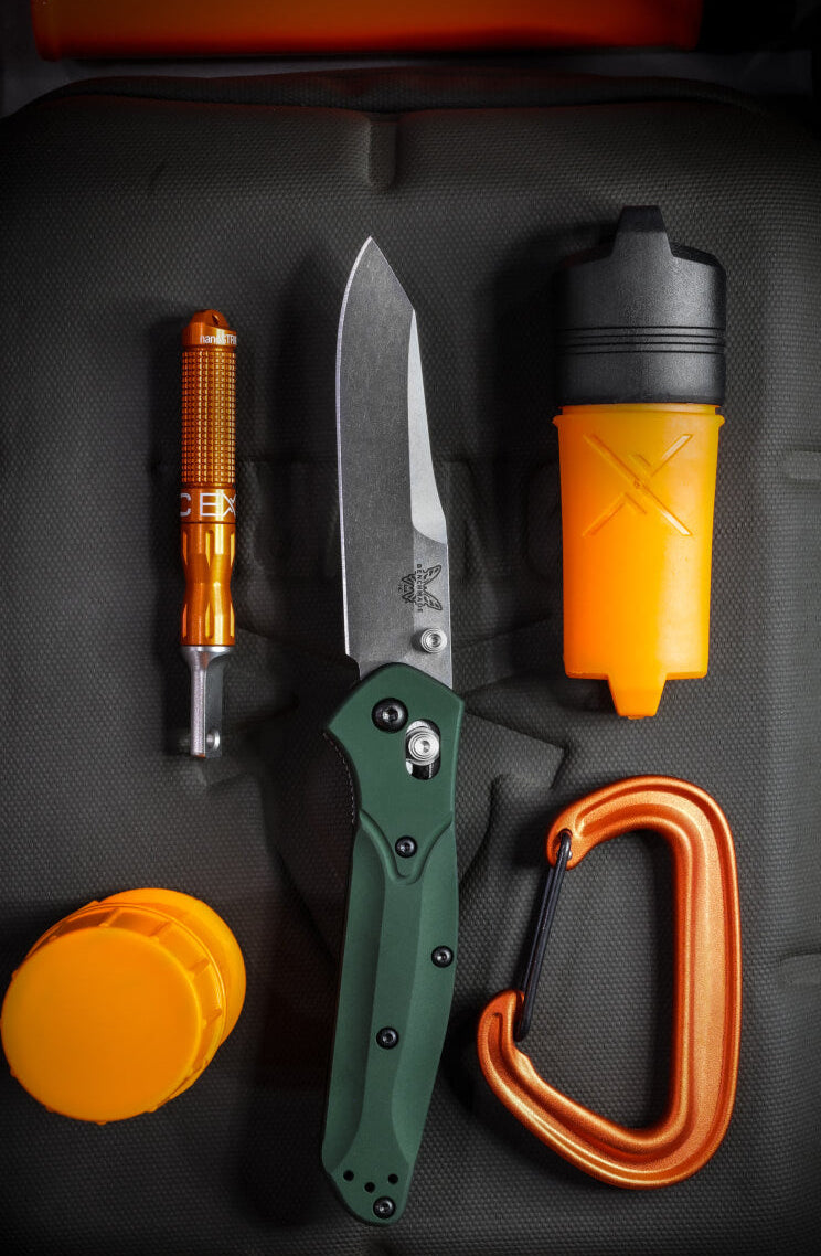 Green folding knife, orange fire starter, carabiner, and containers arranged on dark textured surface.