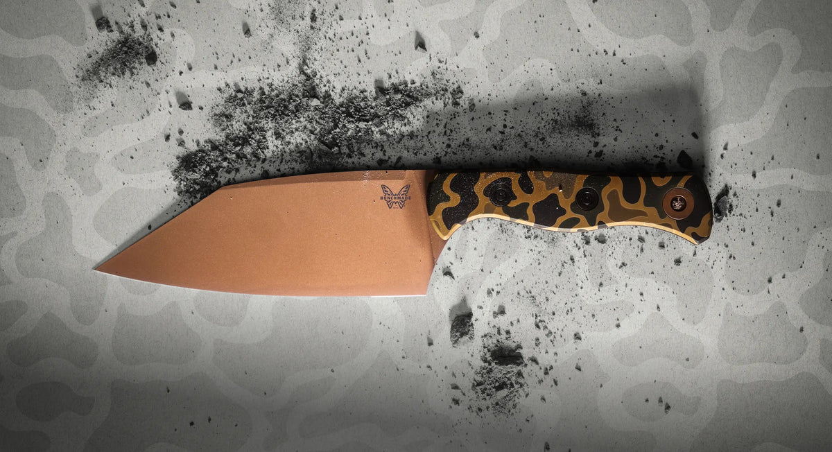 Fixed-blade knife with copper-colored blade and camouflage handle rests on gray camo surface scattered with dark powder.