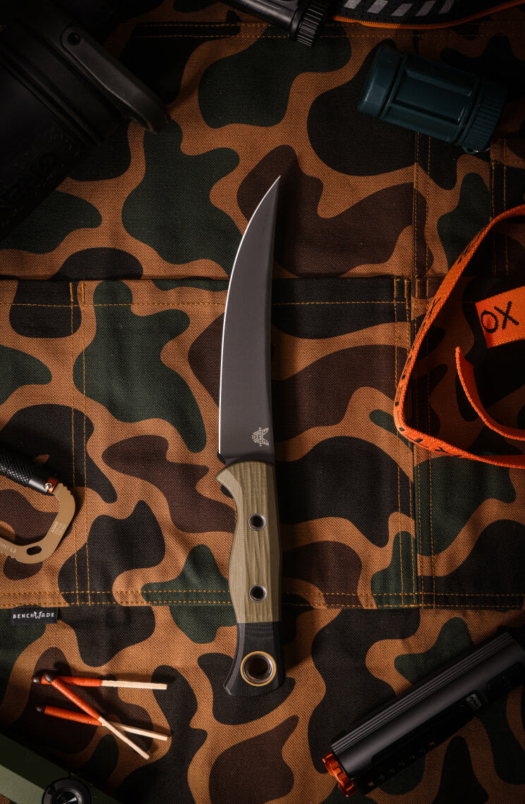 Curved knife with wood handle on camo fabric, surrounded by orange strap, matches, flashlight, and Benchmade tag.