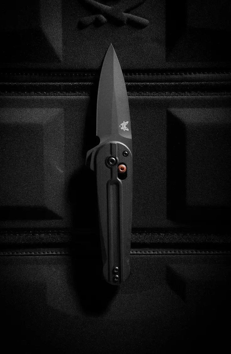 Matte black folding knife with copper pivot rests vertically on a textured black tactical bag in low-key lighting.