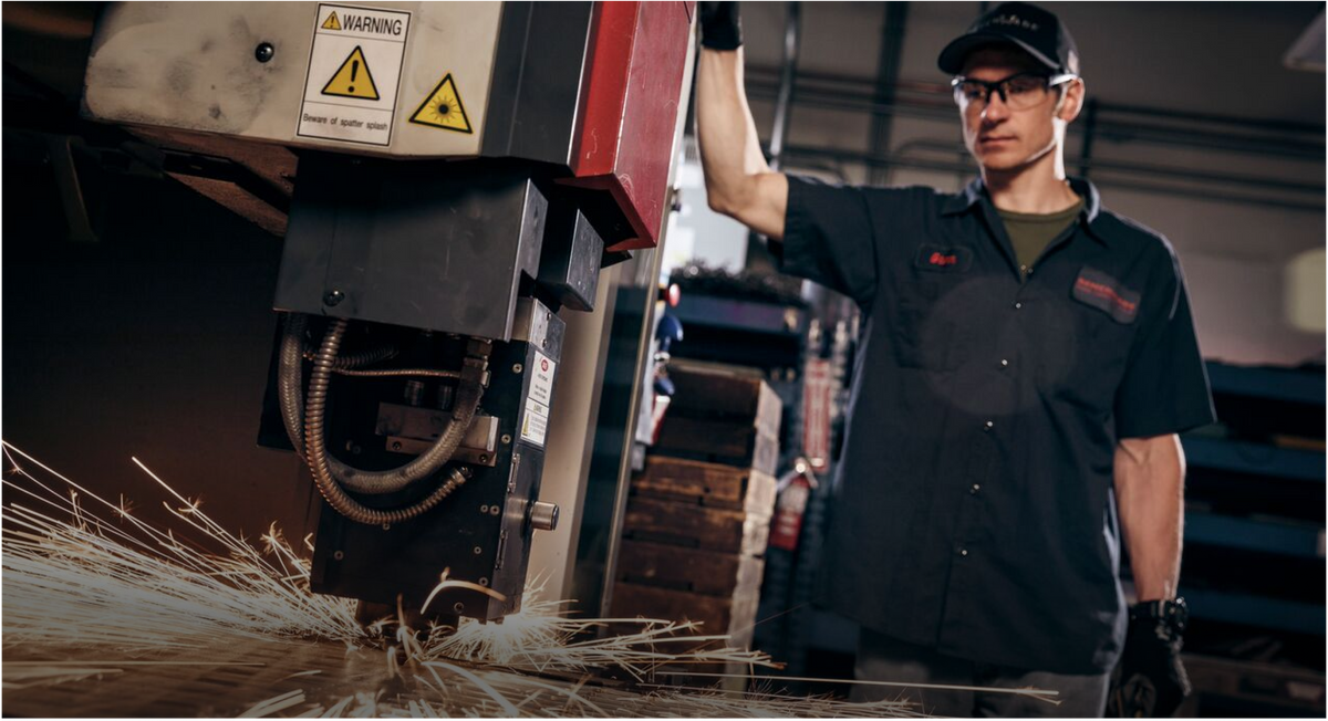 Worker in safety glasses watches laser cutter emit sparks. Yellow warning label visible on machine head.