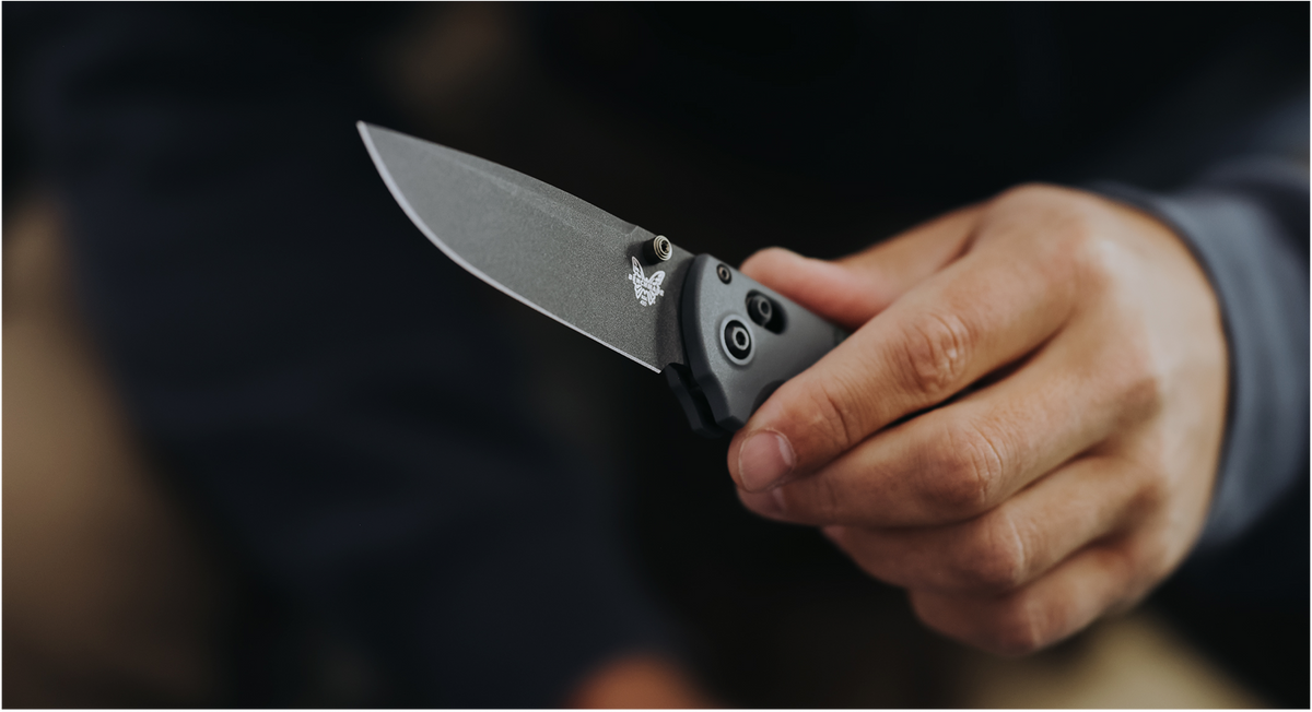 Hand holds open gray folding knife with butterfly logo against dark, blurred background.
