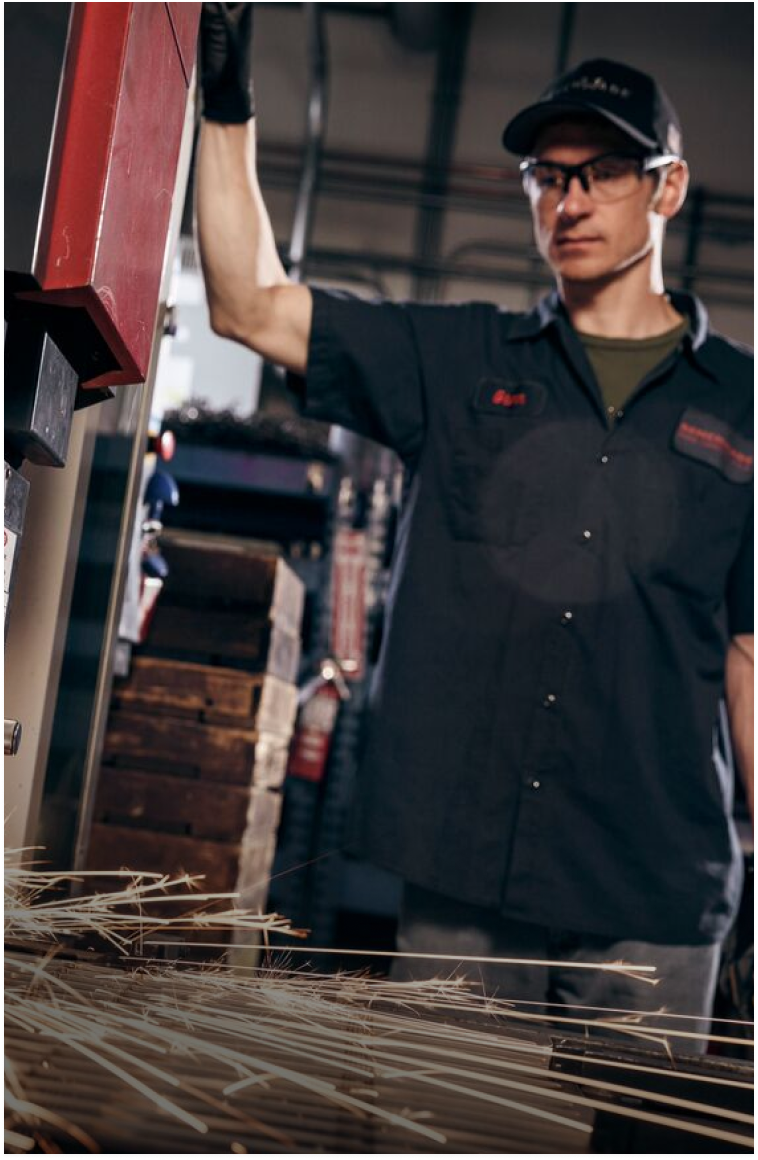 Worker in black shirt, cap, and safety glasses stands by machine as metal shavings fly across foreground.
