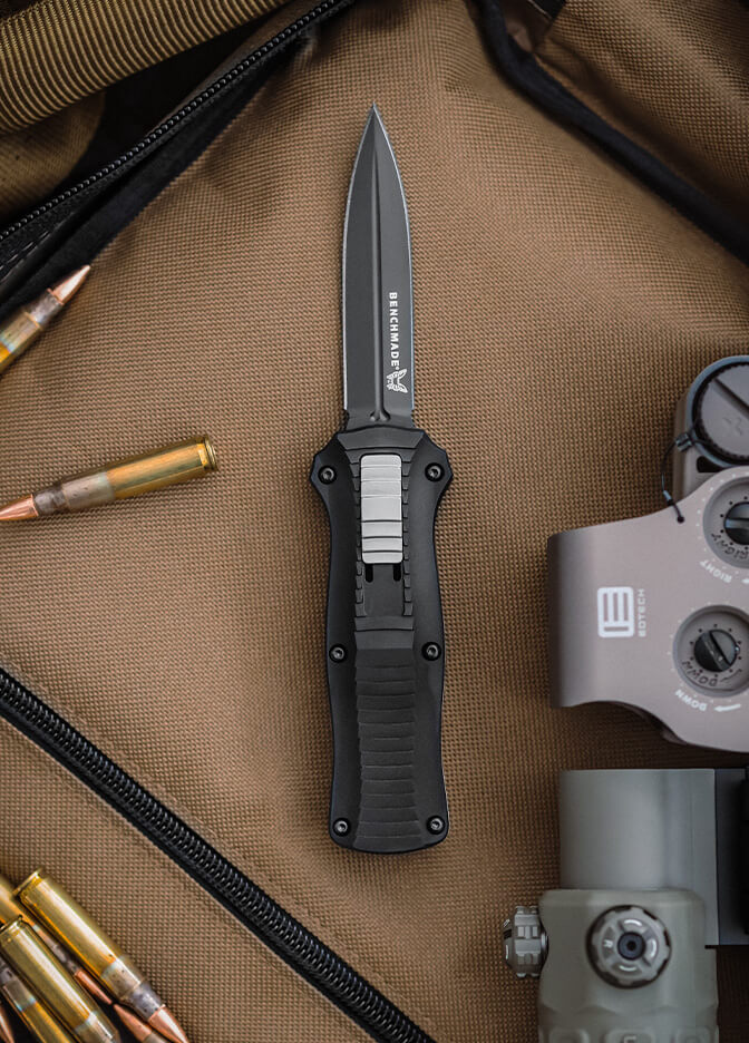 Open Benchmade folding knife on tan canvas, surrounded by brass bullets and a gray tactical scope.