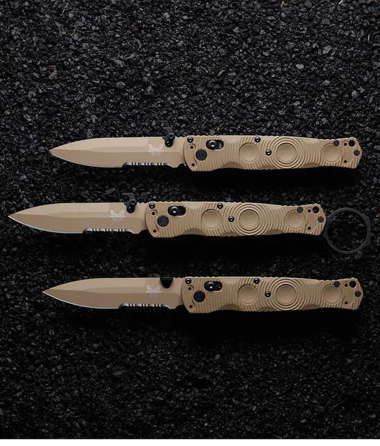 Three tan and black tactical folding knives with butterfly logos, stacked on a dark gravel surface.
