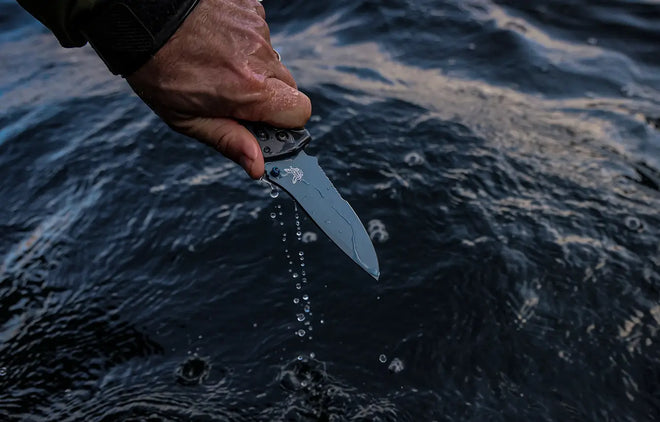 Hand holds wet folding knife over dark rippling water, droplets falling from blade against deep blue surface.