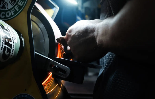 A hand grinds metal on a wheel, creating bright orange sparks in a dark workshop setting.