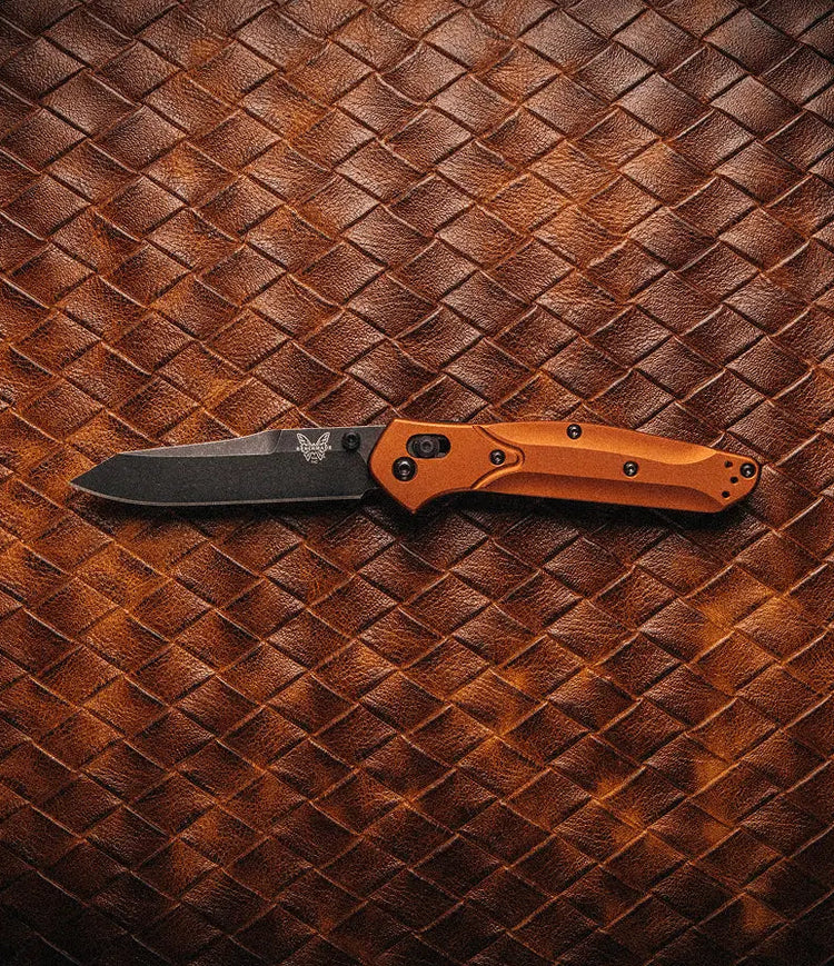 Orange-handled folding knife with black blade and butterfly logo rests on brown woven leather.