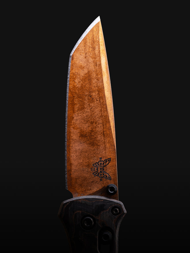 Close-up of a Benchmade folding knife with a bronze, textured blade and dark carbon fiber handle on a black background.