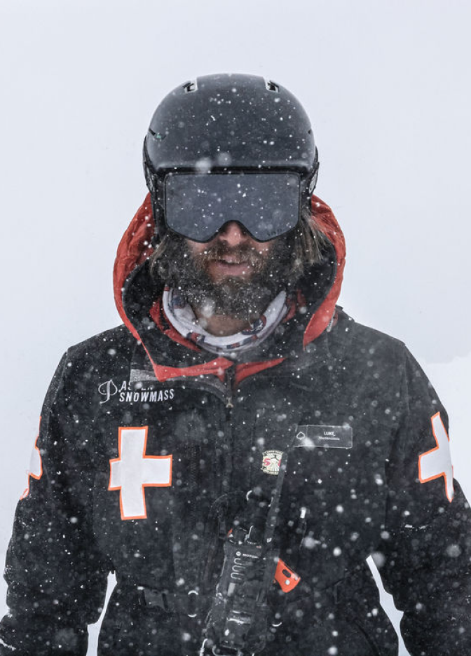 Bearded man in black helmet, goggles, and jacket with white crosses stands outdoors as snow falls around him.