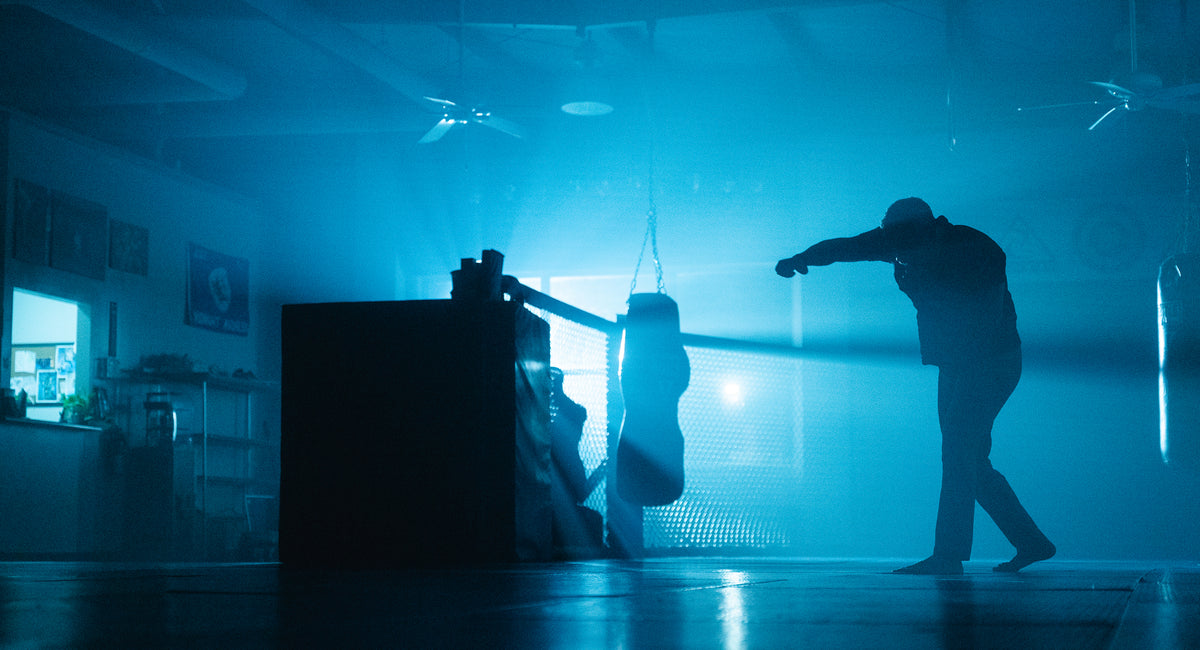 Silhouette of barefoot man punching heavy bag in hazy, blue-lit gym.