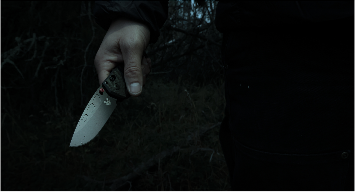 Person's hand grips folding knife in dark, eerie outdoor setting with visible blood droplets on blade.