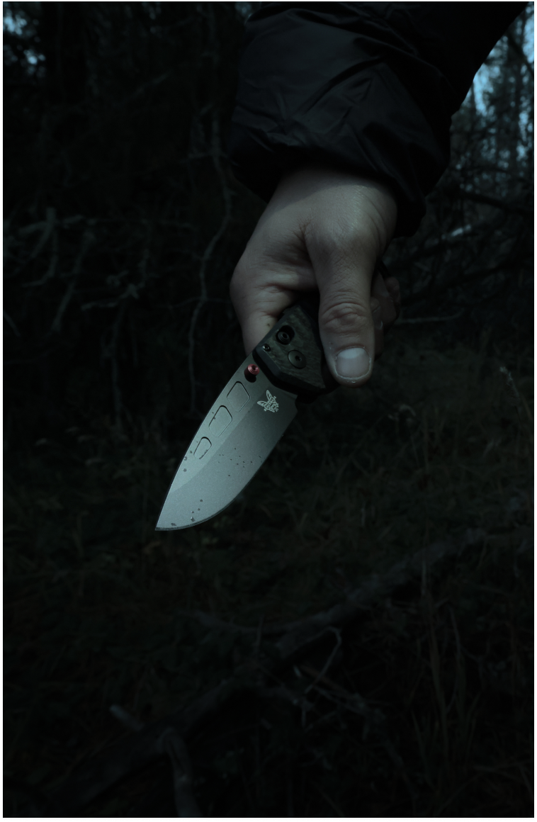 Hand in black sleeve grips folding knife with silver blade and red pivot, set against dark forest background.