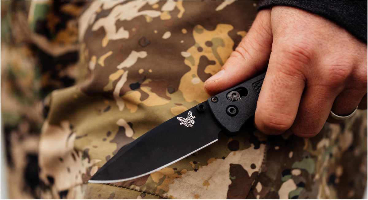 Hand holding black Benchmade folding knife over camouflage fabric.