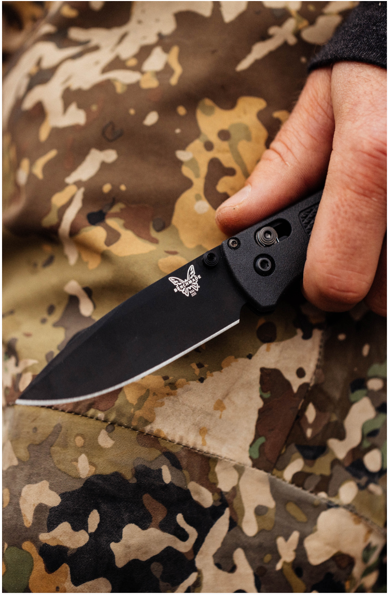 Hand grips black Benchmade folding knife over camouflage fabric, blade open with visible logo.