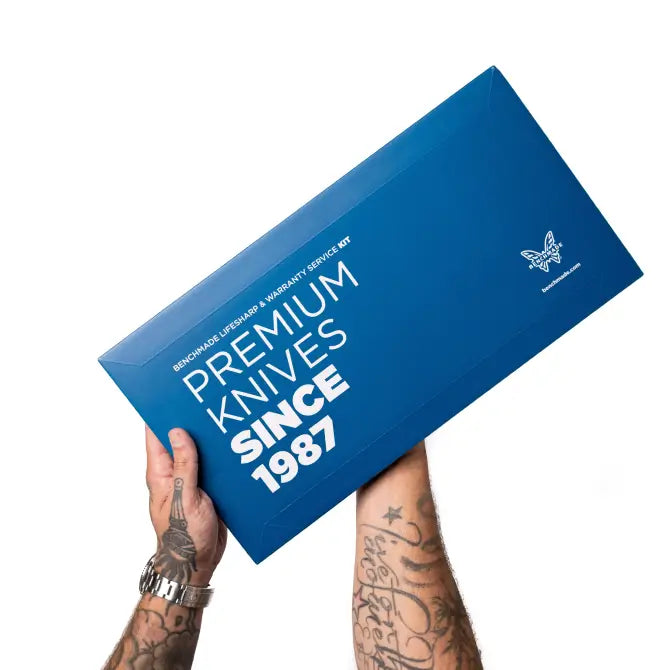 Tattooed hands hold a blue Benchmade box reading "Premium Knives Since 1987" against a white background.