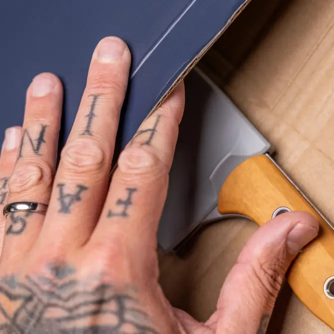 Tattooed hand with silver ring opens blue box, revealing a knife with a light wooden handle inside.