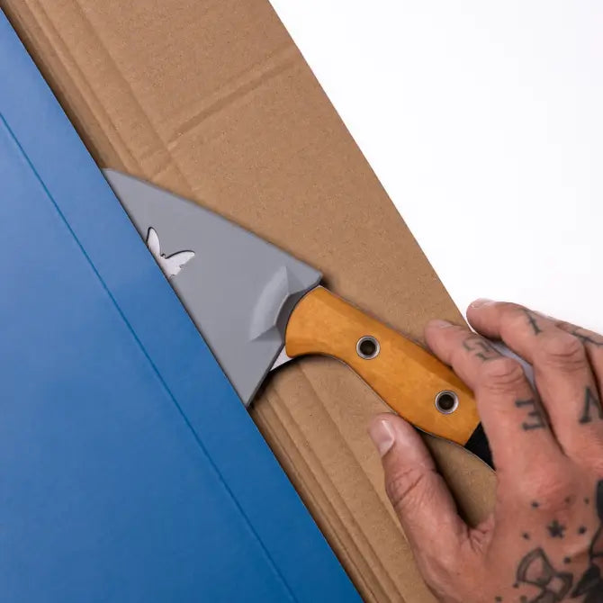 Tattooed hand grips orange-handled knife, cutting blue packaging on brown cardboard against white background.