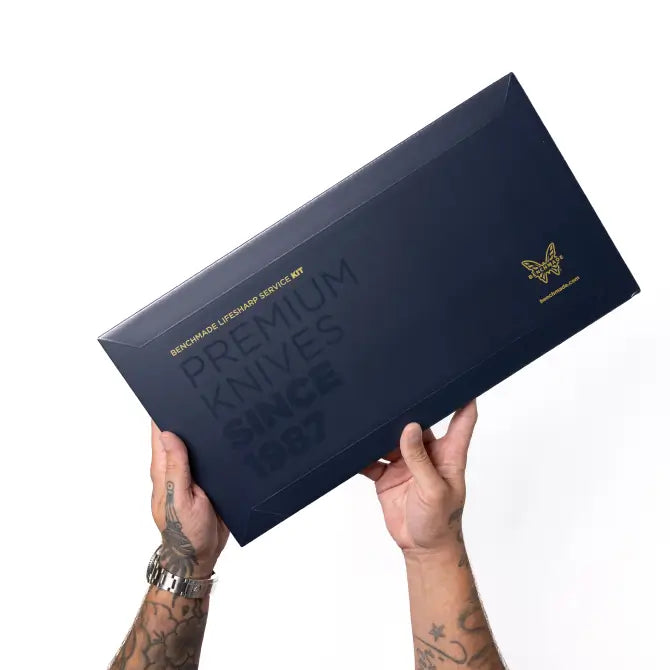Tattooed hands hold a dark blue Benchmade box reading Premium Knives Since 1987 with gold logo.