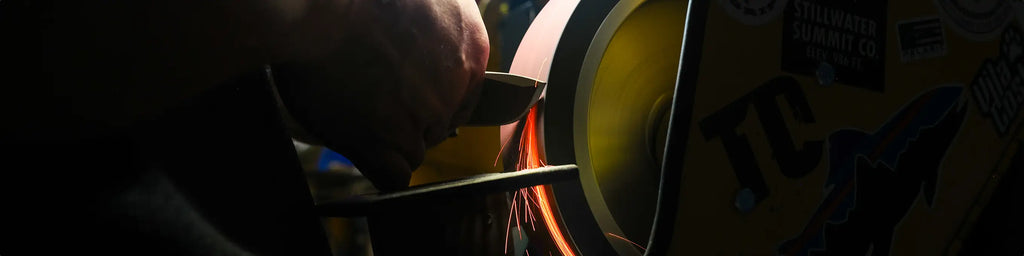 Hand holds metal to grinding wheel, sending bright orange sparks flying in a dimly lit workshop.