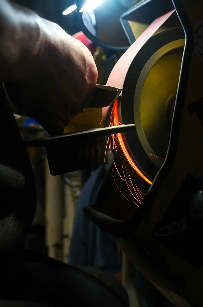 Hand holds metal against spinning belt sander, sending bright orange sparks flying in dimly lit workshop.