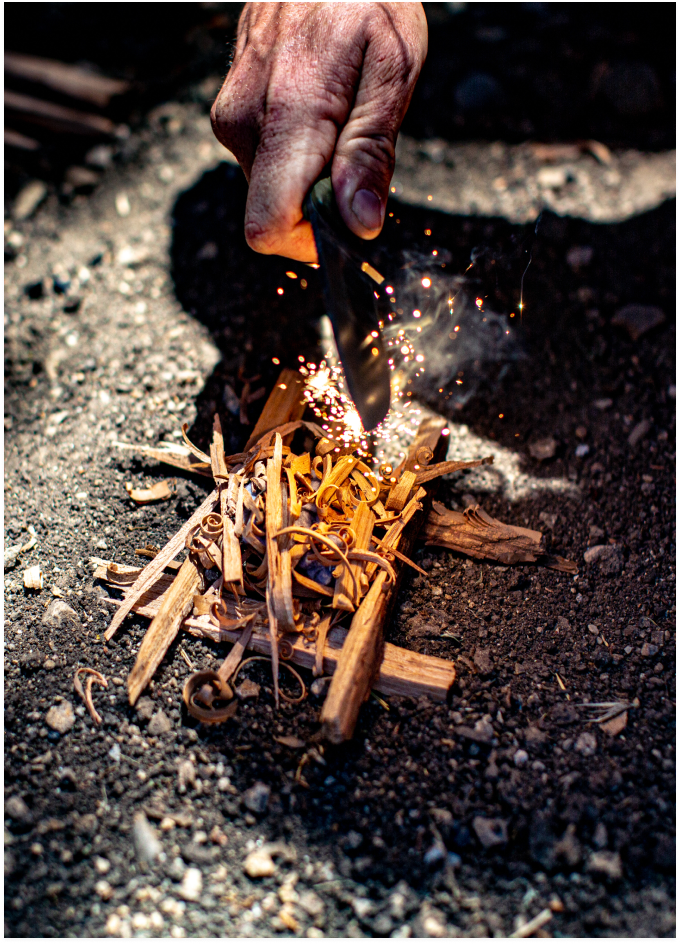 Weathered hand strikes ferro rod over wood shavings, sparking flame on gravel ground in outdoor survival scene.