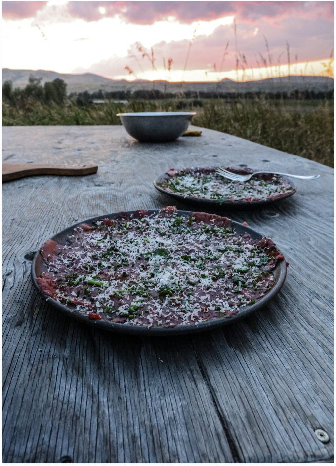 Two plates of carpaccio with herbs and cheese on a wooden table, sunset over mountains in background.