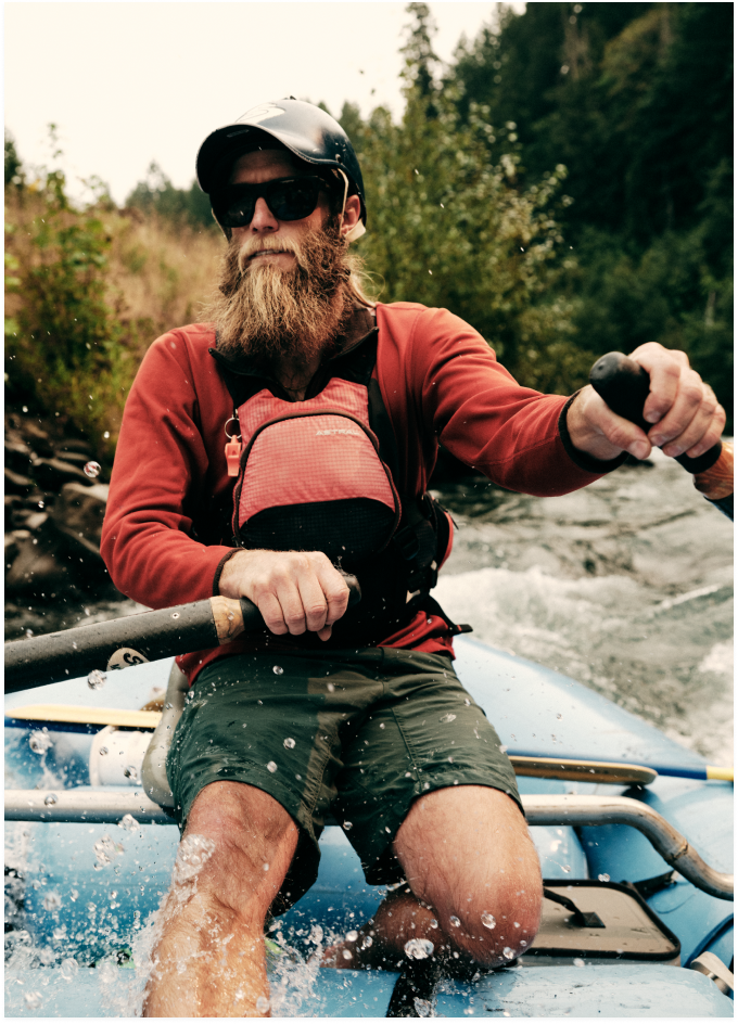 Bearded man in helmet and sunglasses paddles a blue raft through splashing whitewater rapids near forested banks.
