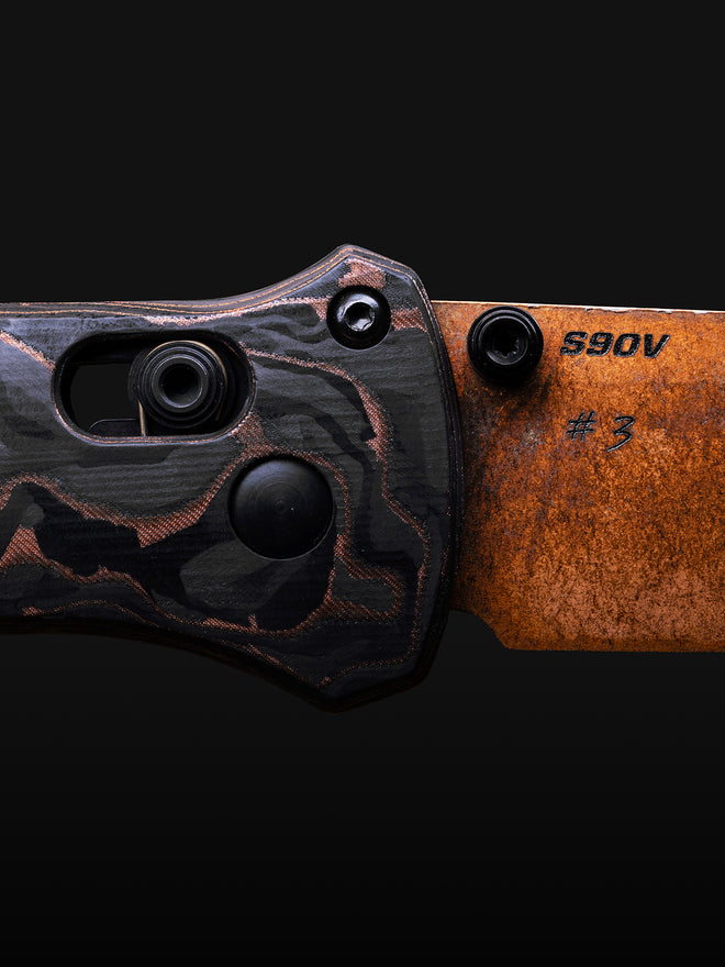 Close-up of a folding knife with black and copper camouflage handle and rusted orange blade engraved "S90V #3".