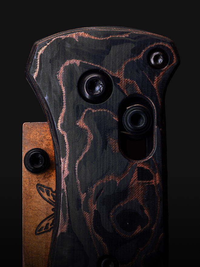 Close-up of a folding knife handle with black and copper marbled scales, showing screws and an engraved butterfly on the pocket clip.