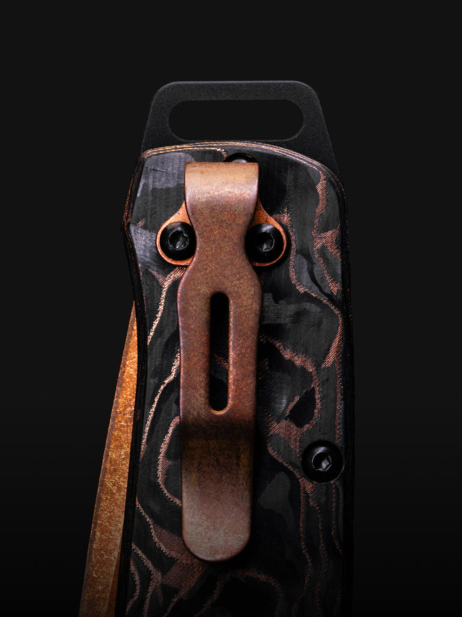 Close-up of a knife handle with black and copper camouflage pattern, featuring a bronze pocket clip against a dark background.