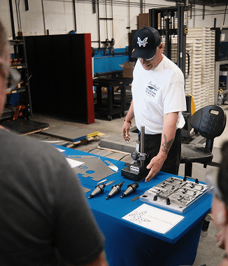 Man in white shirt and cap demonstrates tools on a blue table in an industrial workshop setting.
