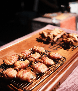 Golden bacon-wrapped items cool on wire racks atop a wooden board, with more cooked pieces in the blurred background.