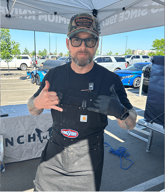 Man in camo hat, sunglasses, and apron gives shaka sign under white canopy at outdoor event.