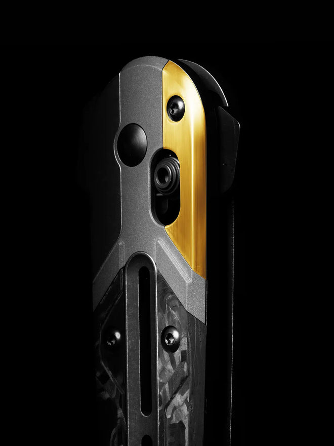 Close-up of a folding knife handle with gray metal, gold inlay, carbon fiber scales, and pocket clip against black background.