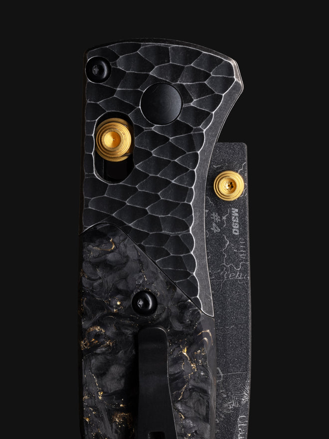 Close-up of folding knife handle with hammered metal, gold inlay, black carbon fiber, and M390 blade marking.
