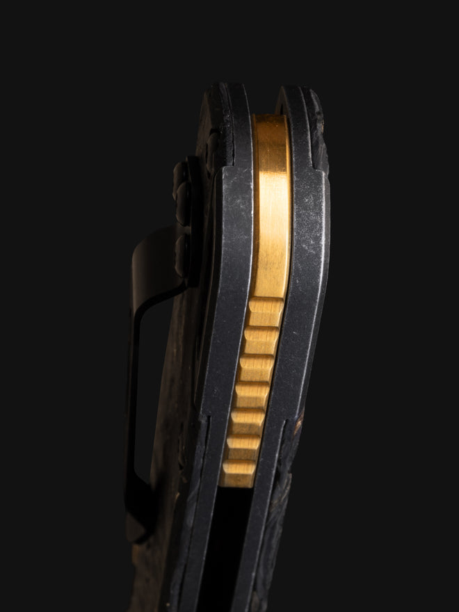 Side profile of a black folding knife with a brass liner lock and pocket clip against a dark background.