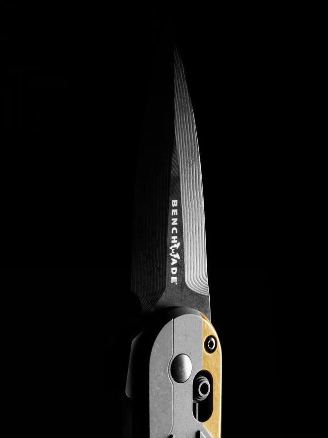 Close-up of a Benchmade folding knife with patterned steel blade and brass handle against a black background.