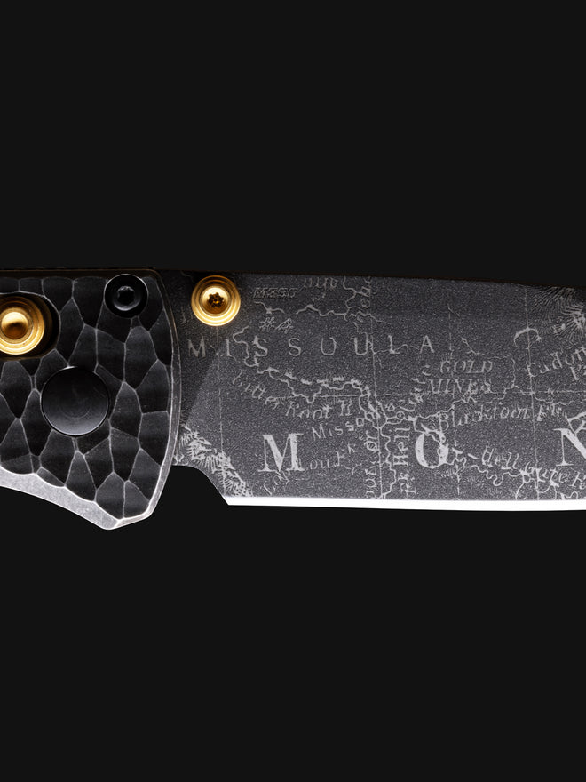 Close-up of a knife blade engraved with "MISSOULA" map and text, featuring a textured black handle with gold screws.
