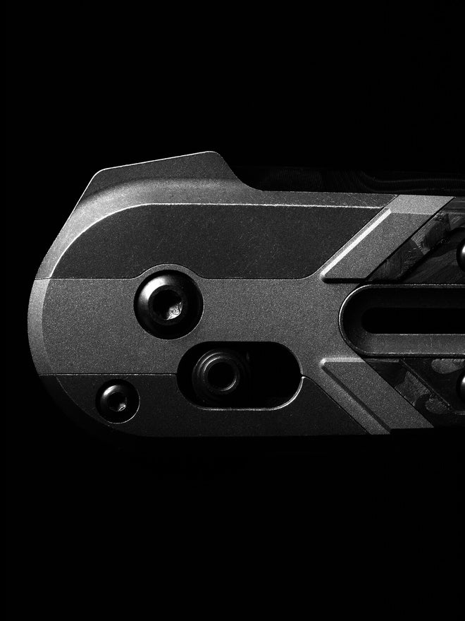 Close-up of a matte black tactical folding knife handle with screws and carbon fiber inlay on a dark background.